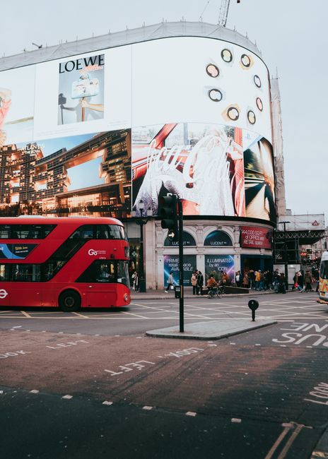 Nighttime scene in Piccadilly Circus showing a red double-decker bus in the foreground at a busy intersection, with illuminated digital billboards displaying advertisements and images on surrounding b