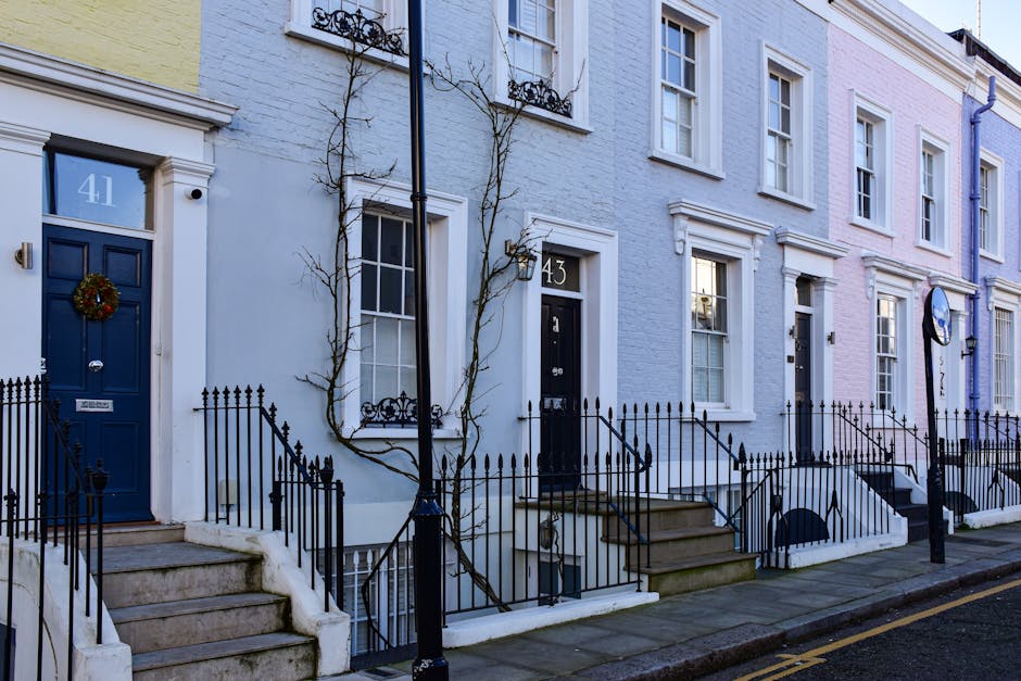 A row of elegant Victorian-style terraced buildings with cream-colored facades, decorative cornices, and large sash windows, situated on a city street with parked cars in front. The buildings feature 