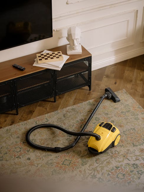 A yellow cylinder-shaped vacuum cleaner with a black hose and handle is situated on a patterned area rug in a room with wooden flooring. Behind it, a black metal media console with a wooden top holds 